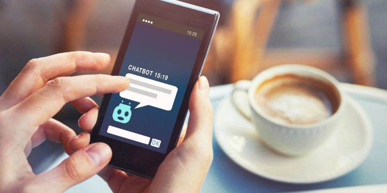 Person am Tisch mit Kaffee tippt auf dem Smartphone in einem Chatbot-Fenster 